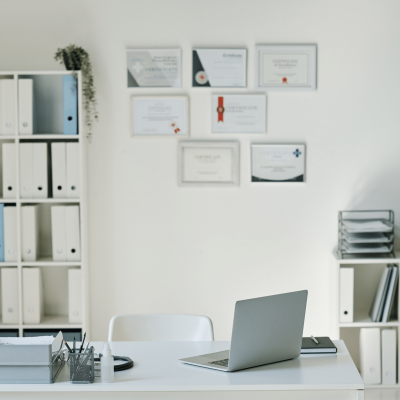 Office desk with a silver laptop, notebook, and pen holder on white desk; blurred certificates on the wall in the background.