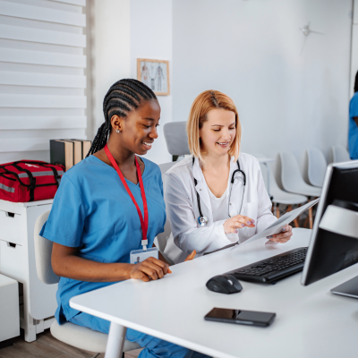 Two healthcare workers—nurse in blue scrubs and doctor in white coat with stethoscope—review documents at a desk with a computer in a bright clinic setting.