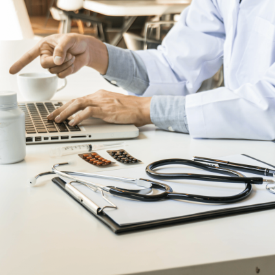 Medical professional in a white coat typing on a laptop at a clinic desk, with a stethoscope, pills, and a syringe visible on the table.